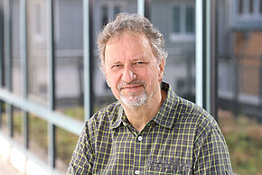 Mann mit grauem Bart und kurzen Haaren, trägt ein kariertes Hemd. Er lächelt freundlich vor einer Fensterfront.