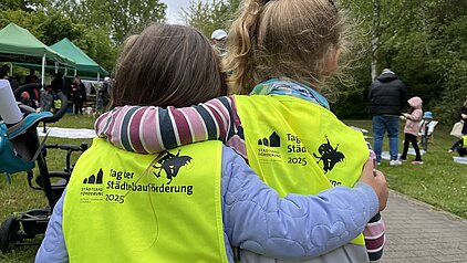 Zwei Kinder mit gelben Warnwesten, die 'Tag der Stadtbauförderung 2025' tragen, umarmen sich auf einer Veranstaltung im Freien