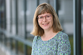Frau mit Brille und bunter Bluse lächelt vor einer Fensterfront.