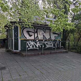 Kleines Gebäude mit Graffiti an den Wänden, umgeben von Bäumen und gepflastertem Platz
