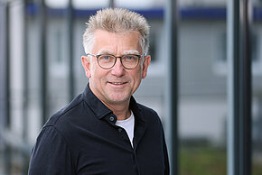 Mann mit grauen Haaren und Brille, trägt ein schwarzes Hemd.