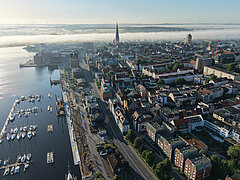 Luftaufnahme eines Stadthafens mit Segelbooten und angrenzender Innenstadt mit Kirchturm und Wohngebäuden unter leichtem Nebel