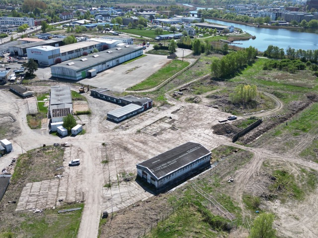 Luftaufnahme des unbebauten Gebietes mit mehreren Lagerhallen, unbefestigten Flächen und der Unterwarnow im Hintergrund