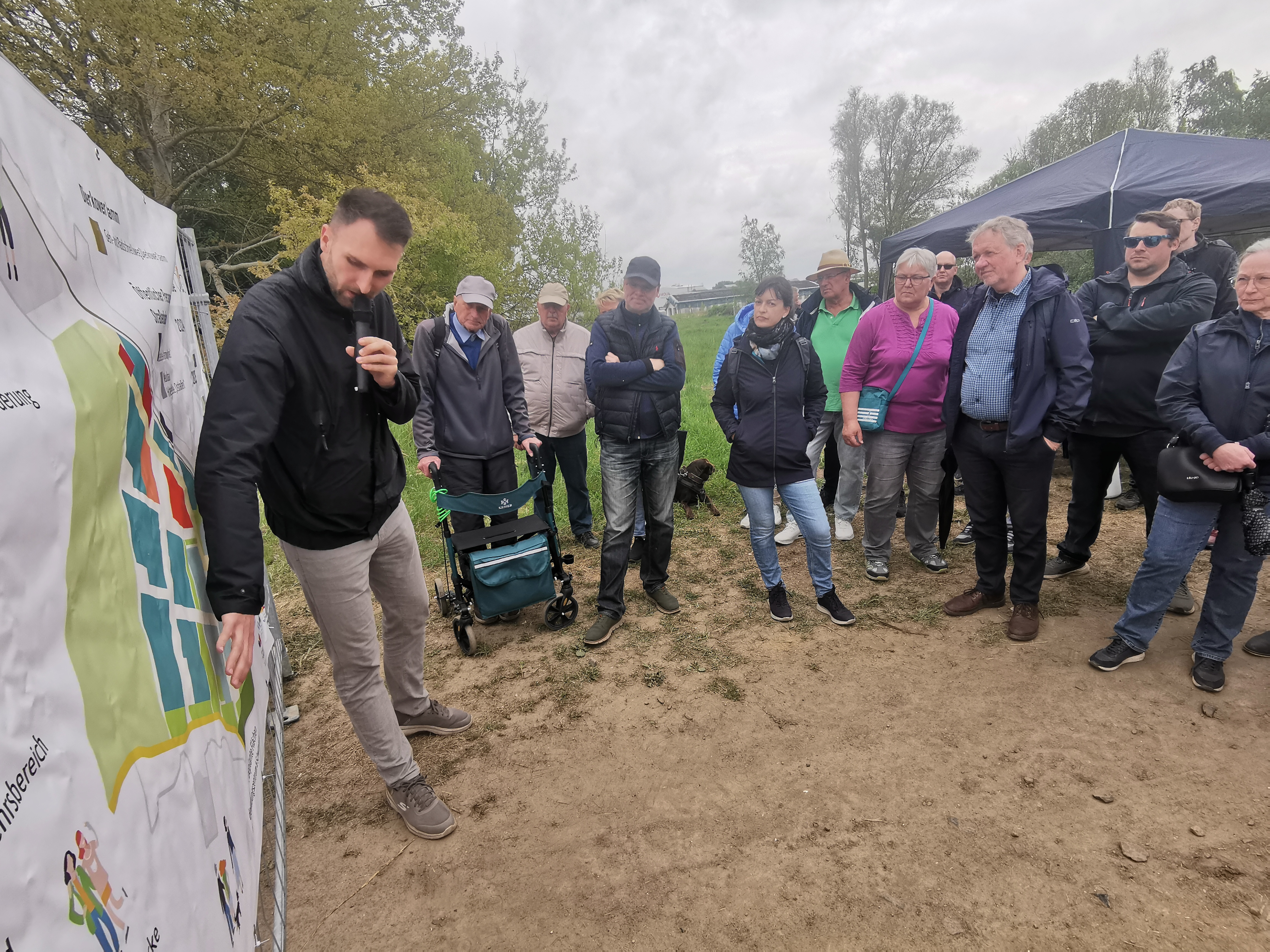 Gruppe von Menschen, die im Freien vor einem Plakat stehen. Ein MAnn hält ein Mirkofon und erklärt und zeigt etwas auf dem Plakat.