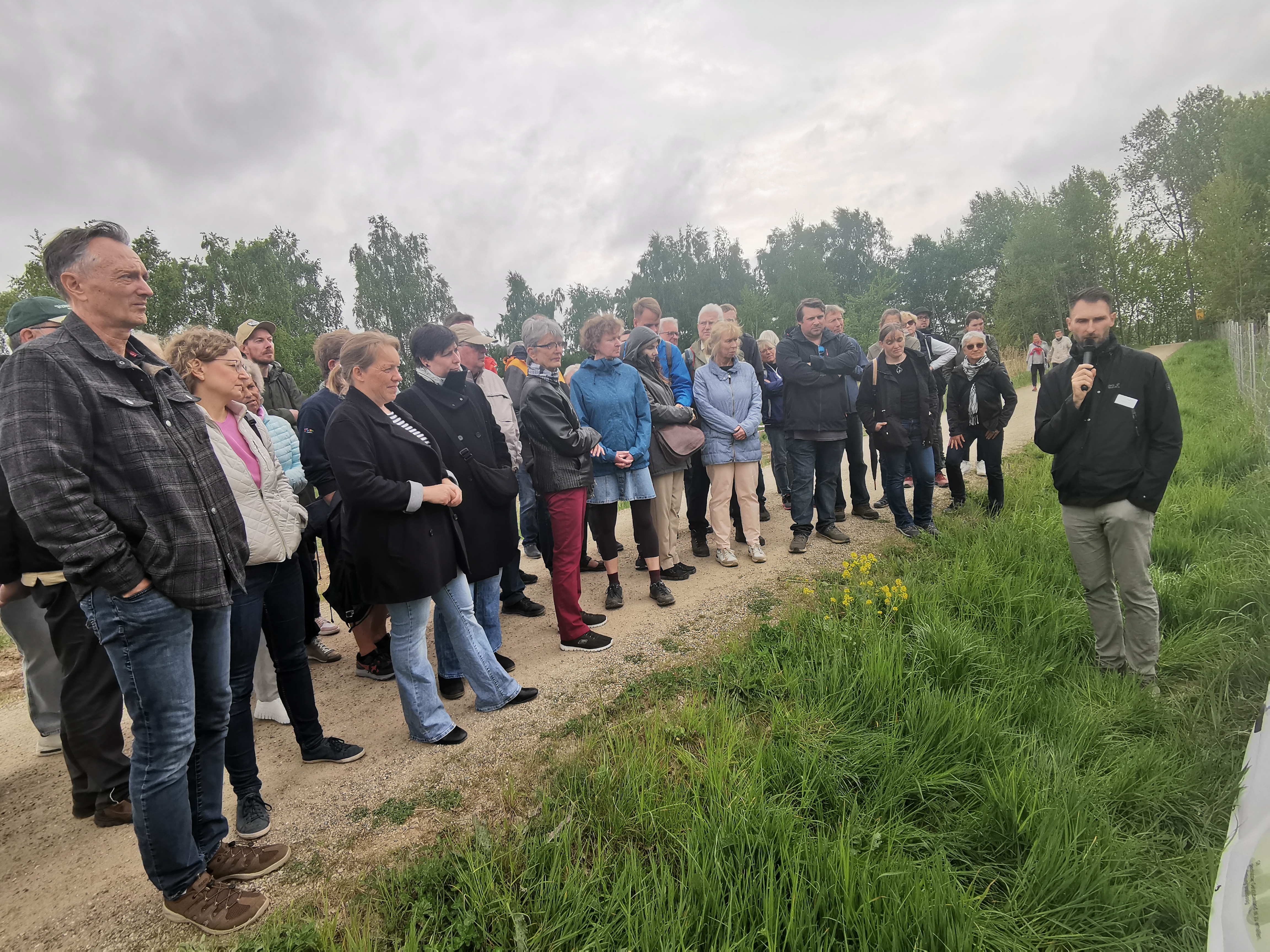 Gruppe von Menschen, die im Freien vor einem Plakat stehen. Ein MAnn hält ein Mirkofon und erklärt etwas auf dem Plakat.