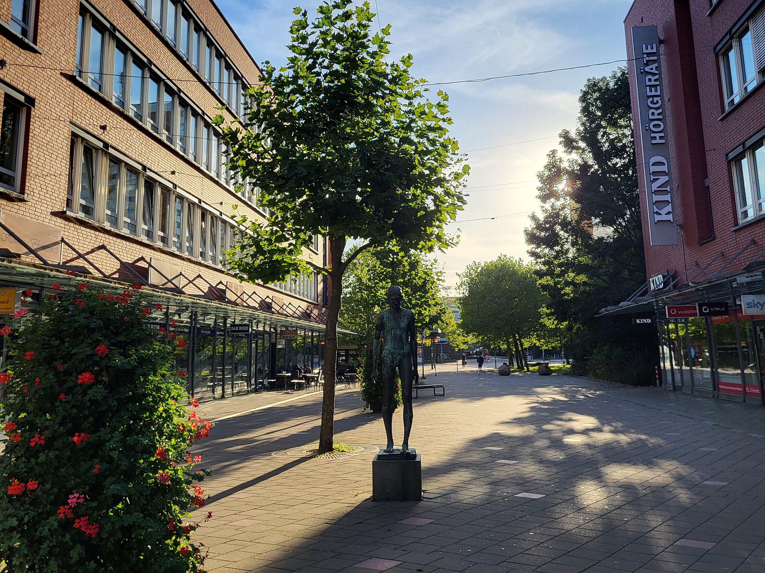 Straßenansicht mit einer Bronzestatue auf einem Sockel, Bäumen und Gebäuden mit Schaufenstern unter blauem Himmel