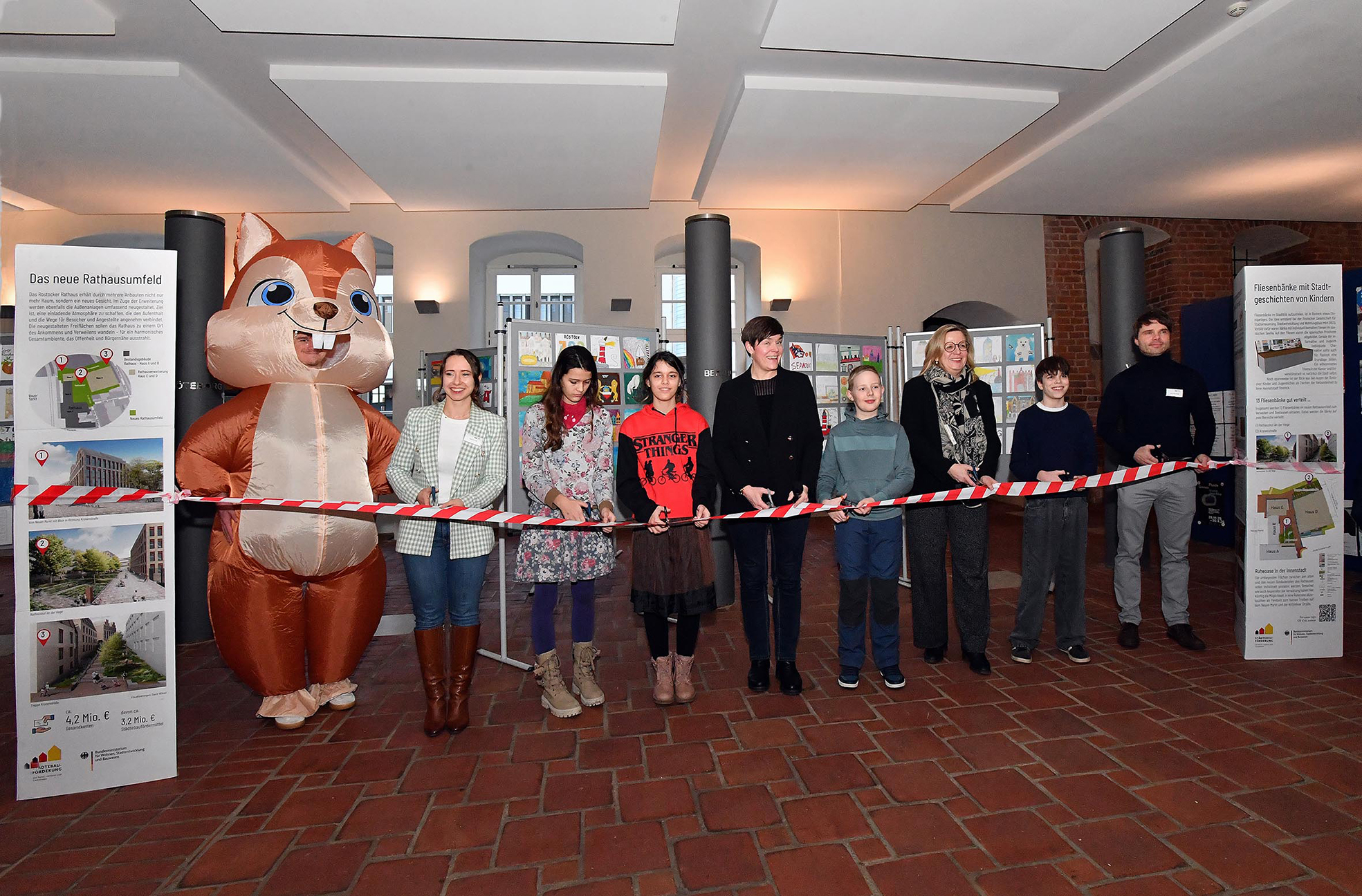 Mehrere Personen stehen in einer Reihe hinter einem rot-weiß gestreiften Absperrband in einem Raum mit Backsteinboden und Informationsständen, links eine Person im Eichhörnchen-Kostüm